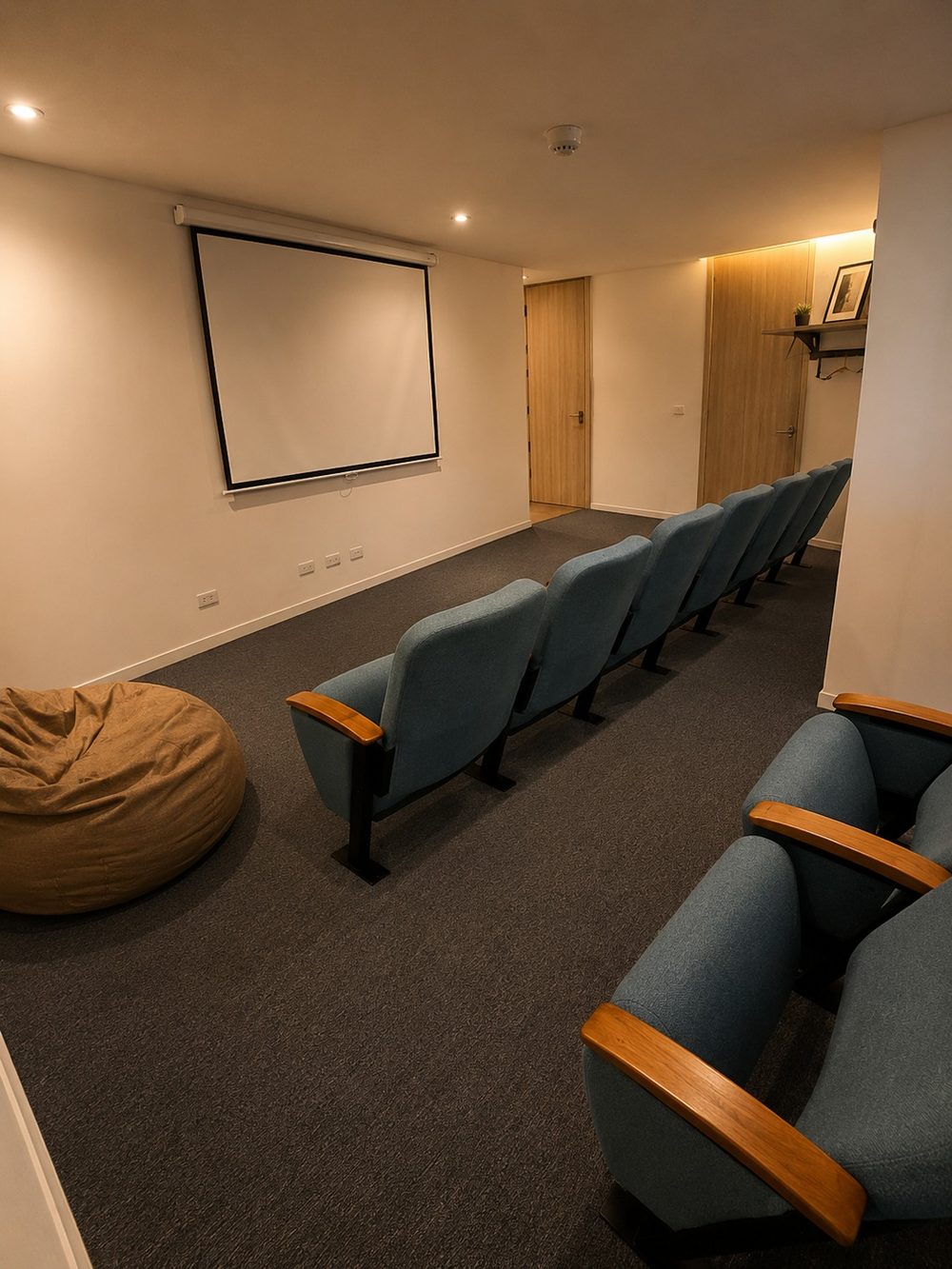Sala de conferencias con dos filas de butacas azul grisáceo frente a una pantalla de proyección desplegada, y un puf en el suelo.